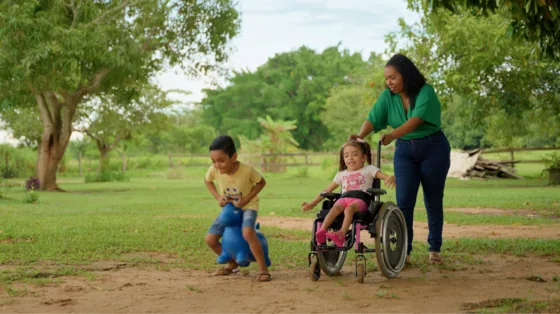 Mãe empurra a cadeira de rodas da filha em um parque, ao lado de outra criança