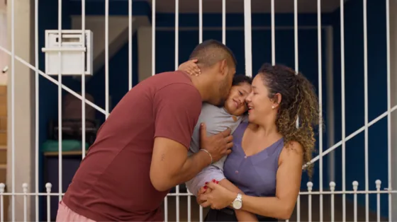 Pai e mãe abraçam o filho pequeno no portão de casa
